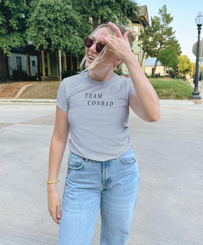 May include: A light gray t-shirt with the text "TEAM CONRAD" in black. The person is wearing light blue jeans and sunglasses. A gold bracelet is visible on the left wrist.