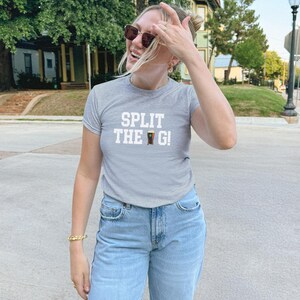 Pu&ograve; includere: Una maglietta grigio chiaro con la scritta bianca "SPLIT THE G!" e due bicchieri di birra marrone scuro. La persona indossa jeans azzurri e occhiali da sole.