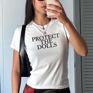 Puede incluir: Camiseta blanca con el texto "PROTECT THE DOLLS" en letras negras. La camiseta tiene mangas cortas y se lleva con un bolso negro. La persona lleva gafas de sol y sostiene un teléfono.