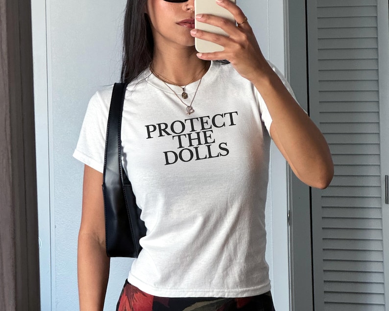 Puede incluir: Camiseta blanca con el texto "PROTECT THE DOLLS" en letras negras. La camiseta tiene mangas cortas y se lleva con un bolso negro. La persona lleva gafas de sol y sostiene un tel&eacute;fono.