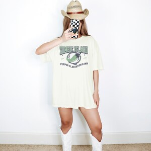May include: A cream colored t-shirt with a graphic print of a green pepper and the text "Zombie Island Pepper Plantation & Inn". The shirt is being worn by a person wearing a straw cowboy hat and white cowboy boots.