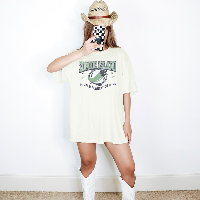 May include: A cream colored t-shirt with a graphic print of a green pepper and the text "Zombie Island Pepper Plantation & Inn". The shirt is being worn by a person wearing a straw cowboy hat and white cowboy boots.