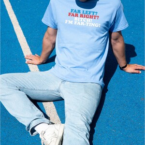 May include: Light blue t-shirt with the text "FAR LEFT? FAR RIGHT? BITCH! I'M FAR-TING!" in red and white. The person is wearing light blue jeans, white sneakers, and a light blue cap. The background is blue.