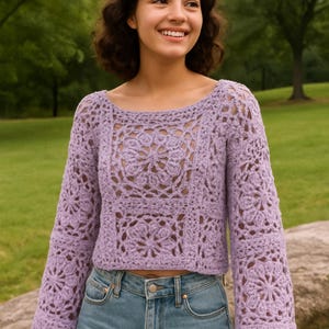 May include: A light purple crochet sweater with a floral pattern. The long-sleeved top has a scoop neck and is paired with blue jeans. The openwork design of the sweater is visible, and the background is a green outdoor setting.