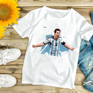 May include: White t-shirt featuring a graphic of a soccer player in an Argentina jersey, number 10, with arms outstretched. The design includes blue accents and a splash effect. The shirt is on a wooden surface.