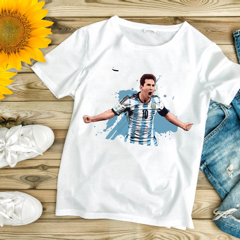 May include: White t-shirt featuring a graphic of a soccer player in an Argentina jersey, number 10, with arms outstretched. The design includes blue accents and a splash effect. The shirt is on a wooden surface.
