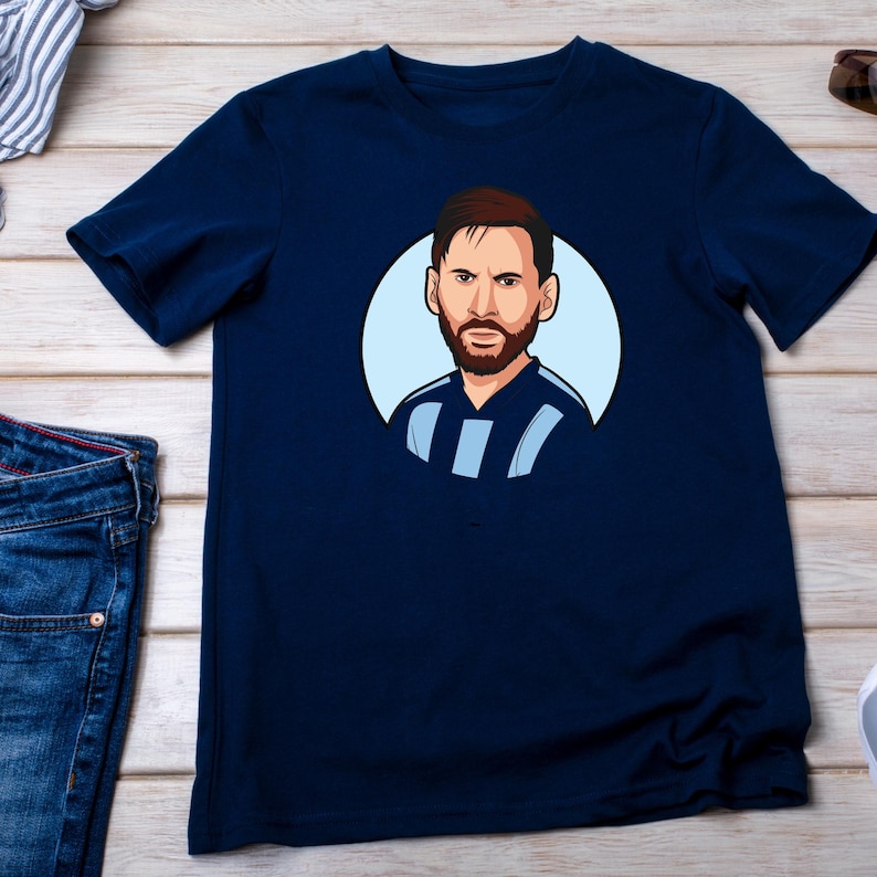 May include: Navy blue t-shirt featuring a cartoon illustration of a man with brown hair and a beard, set within a light blue circle. The man is wearing a blue and white striped shirt. The t-shirt is made of a soft material.