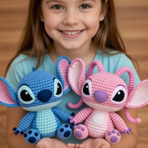 Op de afbeelding: Twee gehaakte pluche speelgoed, een blauwe en een roze, die op stripfiguren lijken. Het blauwe speelgoed heeft grote oren met roze accenten en het roze speelgoed heeft vergelijkbare kenmerken. Beide hebben zwarte ogen en worden door een persoon vastgehouden.