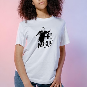 May include: White t-shirt featuring a black graphic of a soccer player in action, with a team crest. The design is centered on the front of the shirt. The shirt is made of a soft material.