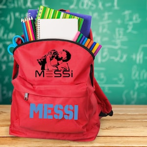 May include: A red backpack filled with school supplies, including pencils, notebooks, and markers. The backpack features a black graphic of a person kissing a trophy, with the name "MESSI" in black and blue.