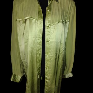 May include: A long-sleeved, button-down shirt in a light green hue. The shirt features a collared neckline, two chest pockets, and satin trim on the cuffs and collar. The fabric has a subtle sheen, and the shirt is displayed against a black background.
