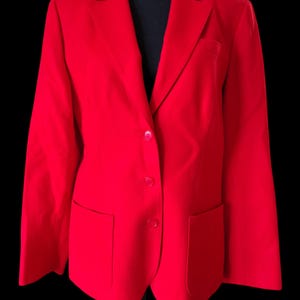 Op de afbeelding: Een felrode blazer met een klassiek ontwerp. De blazer heeft een revers, twee voorzakken en een sluiting met twee knopen. De mouwen zijn lang. Het kledingstuk wordt gepresenteerd tegen een zwarte achtergrond.
