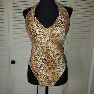 May include: A one-piece swimsuit with a halter top and a leopard print pattern in shades of brown and tan. The swimsuit is displayed on a black mannequin. The fabric has a textured appearance.