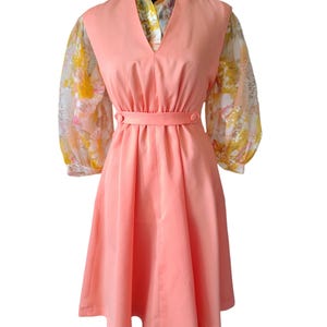May include: A vintage peach-colored dress with a V-neck and a fitted waist. The dress features a peach-colored bodice and a skirt, with sheer, floral-patterned sleeves. The dress has a matching belt and is displayed on a black mannequin.