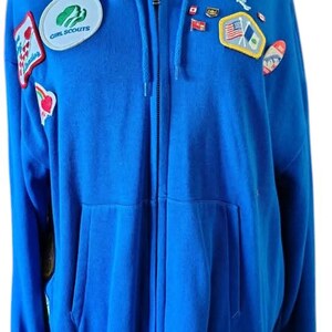 May include: A blue zip-up hoodie with a hood and front pockets. Patches include a Girl Scouts emblem and various flag and emblem patches. The hoodie is made of a soft material.