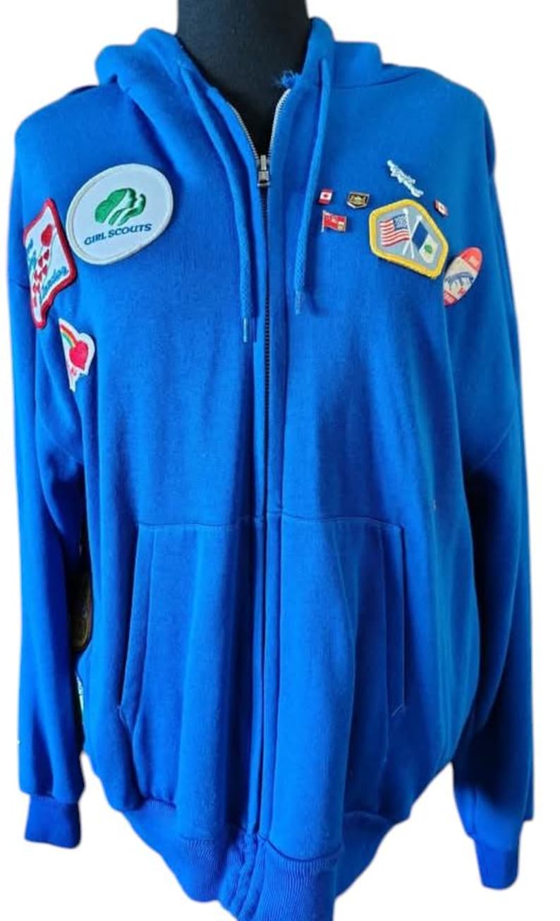 May include: A blue zip-up hoodie with a hood and front pockets. Patches include a Girl Scouts emblem and various flag and emblem patches. The hoodie is made of a soft material.