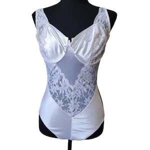 May include: White satin bodysuit with lace detailing. The bodysuit features a sweetheart neckline, lace trim on the straps, and a lace panel down the front. The garment is displayed on a black mannequin.