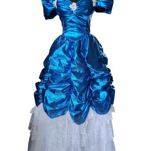May include: A vibrant blue formal dress with a shiny, ruffled bodice and off-the-shoulder sleeves. The dress features white lace detailing at the skirt's hem and small white floral accents. The dress is displayed on a black mannequin.