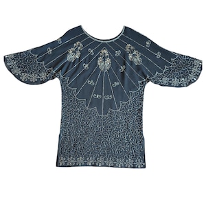 Puede incluir: Top azul oscuro con cuentas y mangas anchas. El top presenta intrincados bordados plateados a lo largo del escote, las mangas y el corpiño. El diseño incluye patrones florales y geométricos, creando un estilo vintage o art déco.