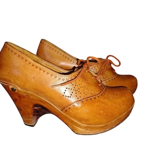 Pode incluir: Um par de tamancos de couro castanho com salto de madeira e sola plataforma. Os sapatos têm um design com atacadores com perfurações decorativas e uma borda ondulada na parte superior.