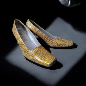 Puede incluir: Un par de zapatos de tacón alto amarillos y texturizados con punta cuadrada. Los zapatos tienen un patrón similar al de un reptil y un tacón pequeño. La marca "BELLINI" es visible dentro del zapato.