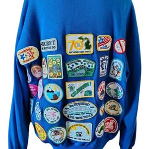 May include: A royal blue hooded sweatshirt adorned with numerous colorful patches. The patches feature text and graphics, including "SECRET SUCCESS 90", "MICHIGAN STATE PARKS", and "GIRL SCOUTS 25th". The sweatshirt has long sleeves and a hood.