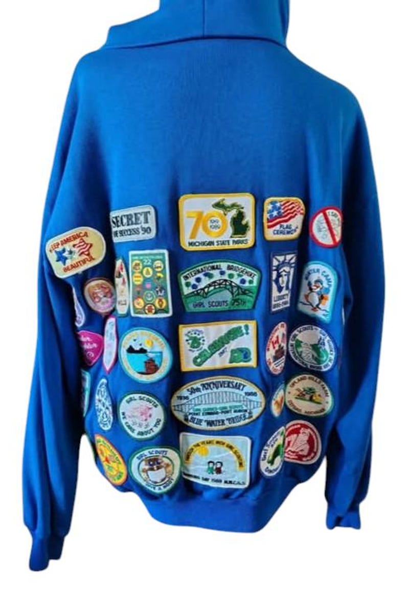 May include: A royal blue hooded sweatshirt adorned with numerous colorful patches. The patches feature text and graphics, including "SECRET SUCCESS 90", "MICHIGAN STATE PARKS", and "GIRL SCOUTS 25th". The sweatshirt has long sleeves and a hood.