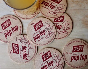 May include: Close-up of six round, brown paper coasters with the Schlitz logo and the text "Pop Top" in red. The coasters feature a hand opening a can of beer.
