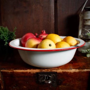 May include: A white enamel bowl with a red rim filled with lemons and pomegranates. The bowl sits on a wooden surface. The lemons are yellow, and the pomegranates are red.