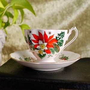 May include: A white teacup and saucer with a Christmas-themed design. The design features red poinsettia flowers, green holly leaves, and pine cones. The cup and saucer have a gold trim.