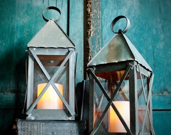 May include: Two antique-style metal lanterns with glass panels and a candle inside. The lanterns have a triangular roof and a handle at the top. They are sitting on a dark book, with a teal-colored wooden background.