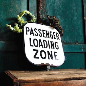 Może przedstawiać: Biały znak „Passenger Loading Zone” z czarnym napisem, zamontowany na drewnie. Znak ma lekko zniszczony wygląd. Zieleń zdobi znak na tle turkusowego drewnianego tła.