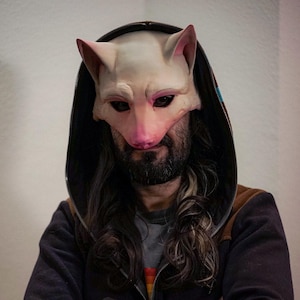 Puede incluir: Una persona con una sudadera con capucha negra con parches de cuero marrón en los codos y una máscara de gato blanca. La máscara tiene detalles rosas alrededor de los ojos y la nariz.
