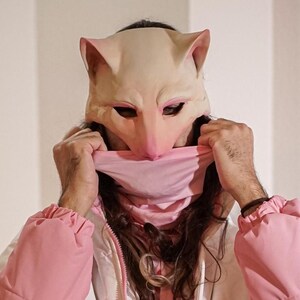 Puede incluir: Una persona lleva una chaqueta acolchada rosa y blanca degradada con una máscara de animal rosa. La persona está de pie frente a una pared con luces de cadena.