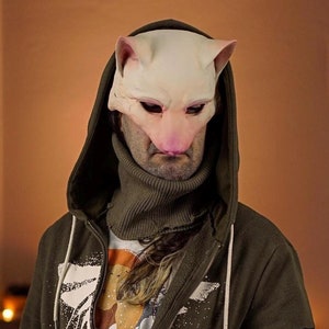 Op de afbeelding: Een persoon draagt ​​een groene hoodie met een witte grafische print en een witte rits. De hoodie heeft een zwarte patch op de linkermouw met een wit libellenontwerp. De persoon draagt ​​ook een blauwe jeans en een witte turtleneck. De persoon draagt ​​een wit kattenmasker met roze oren en neus.