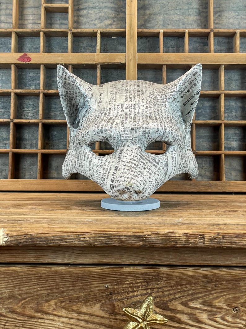 Op de afbeelding: Een handgemaakt vossenmasker van krantenpapier, met een textuuroppervlak en een neutraal kleurenpalet. Het masker is gemonteerd op een lichtblauwe cirkelvormige basis, die de ingewikkelde details van de papierconstructie laat zien. Het masker wordt tentoongesteld op een houten oppervlak.