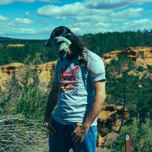 Puede incluir: Una persona con una camiseta azul claro con un dise&ntilde;o gr&aacute;fico, vaqueros azules y un cubrecabezas. El fondo presenta una vista panor&aacute;mica de un bosque y un cielo nublado.