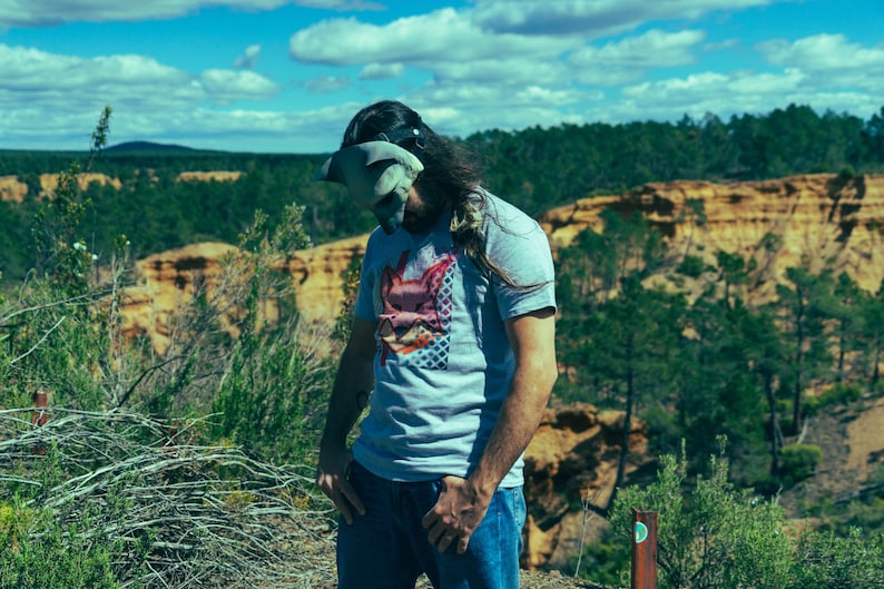 Puede incluir: Una persona con una camiseta azul claro con un dise&ntilde;o gr&aacute;fico, vaqueros azules y un cubrecabezas. El fondo presenta una vista panor&aacute;mica de un bosque y un cielo nublado.
