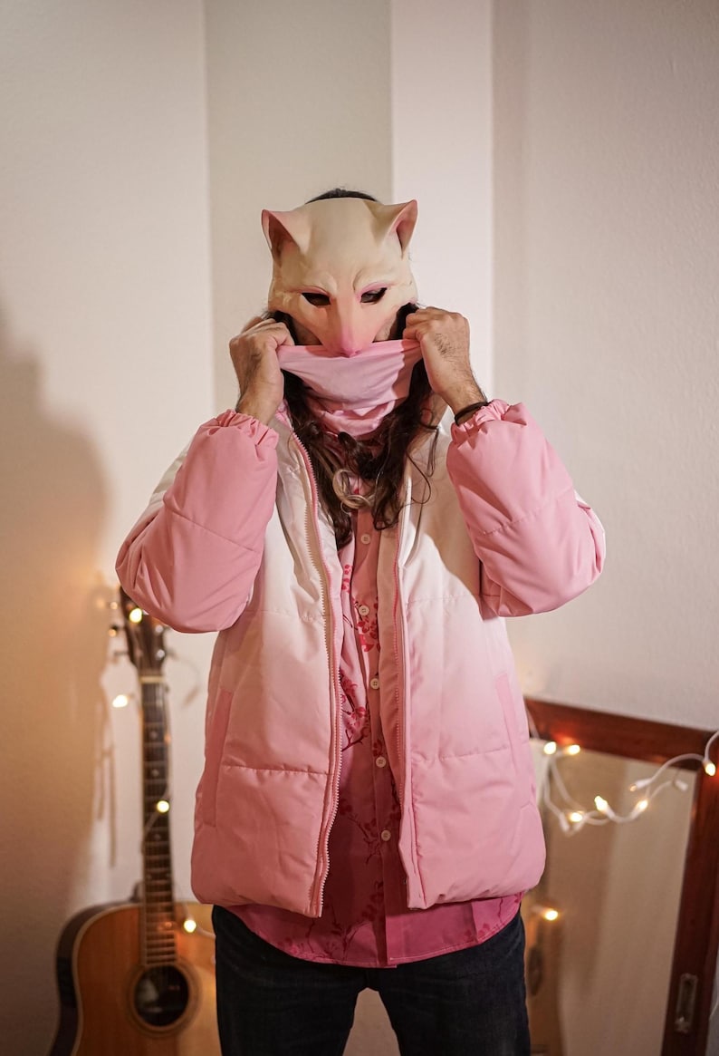 Puede incluir: Una persona lleva una chaqueta acolchada rosa y blanca degradada con una máscara de animal rosa. La persona está de pie frente a una pared con luces de cadena.