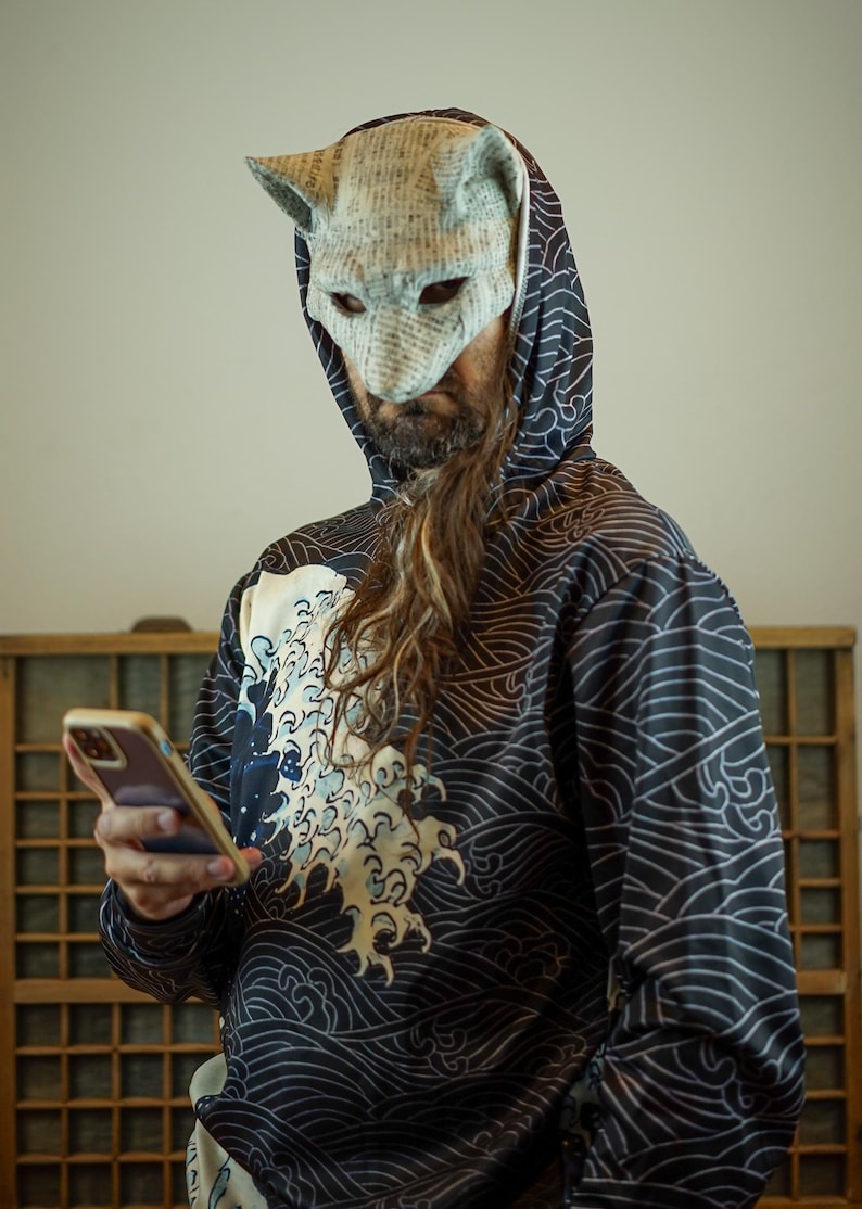 Puede incluir: Una persona con una máscara de gato de papel maché y una sudadera con capucha negra con un patrón de olas. La sudadera tiene un diseño de olas blanco y azul. La persona sostiene un teléfono inteligente.