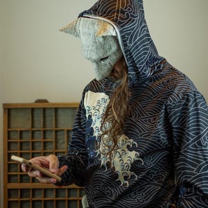 Op de afbeelding: Een zwarte hoodie met ritssluiting met een golfpatroon en een witte en blauwe golfafbeelding. De persoon draagt een papieren masker en houdt een houten telefoonhoesje vast. De hoodie heeft een capuchon.