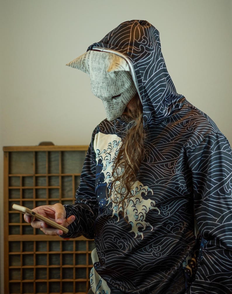 Op de afbeelding: Een zwarte hoodie met ritssluiting met een golfpatroon en een witte en blauwe golfafbeelding. De persoon draagt een papieren masker en houdt een houten telefoonhoesje vast. De hoodie heeft een capuchon.