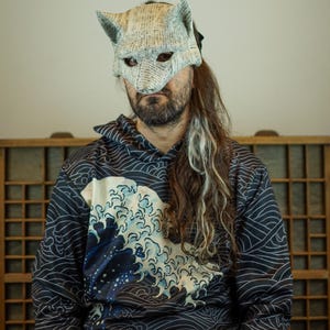 Op de afbeelding: Een handgemaakt wolfsmasker van krantenpapier, gedragen met een marineblauwe en zwarte hoodie met een wit en blauw golfpatroon. Het masker heeft puntige oren en ooggaten. De hoodie heeft een capuchon met trekkoord en lange mouwen.