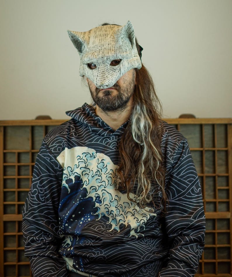 Op de afbeelding: Een handgemaakt wolfsmasker van krantenpapier, gedragen met een marineblauwe en zwarte hoodie met een wit en blauw golfpatroon. Het masker heeft puntige oren en ooggaten. De hoodie heeft een capuchon met trekkoord en lange mouwen.