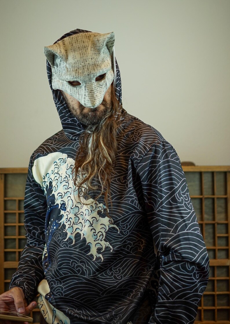 Op de afbeelding: Een persoon draagt een zwarte hoodie met een wit golfpatroon en een papieren masker. Het masker is gemaakt van papier met tekst erop gedrukt. Het ontwerp van de hoodie staat op de voorkant en de mouwen.