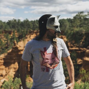 Puede incluir: Una camiseta gris con un gr&aacute;fico de zorro y una m&aacute;scara de zorro. El gr&aacute;fico del zorro es rosa y rojo con un patr&oacute;n geom&eacute;trico. La m&aacute;scara es gris y blanca. El fondo es un paisaje natural.