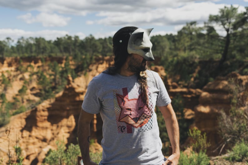 Puede incluir: Una camiseta gris con un gr&aacute;fico de zorro y una m&aacute;scara de zorro. El gr&aacute;fico del zorro es rosa y rojo con un patr&oacute;n geom&eacute;trico. La m&aacute;scara es gris y blanca. El fondo es un paisaje natural.