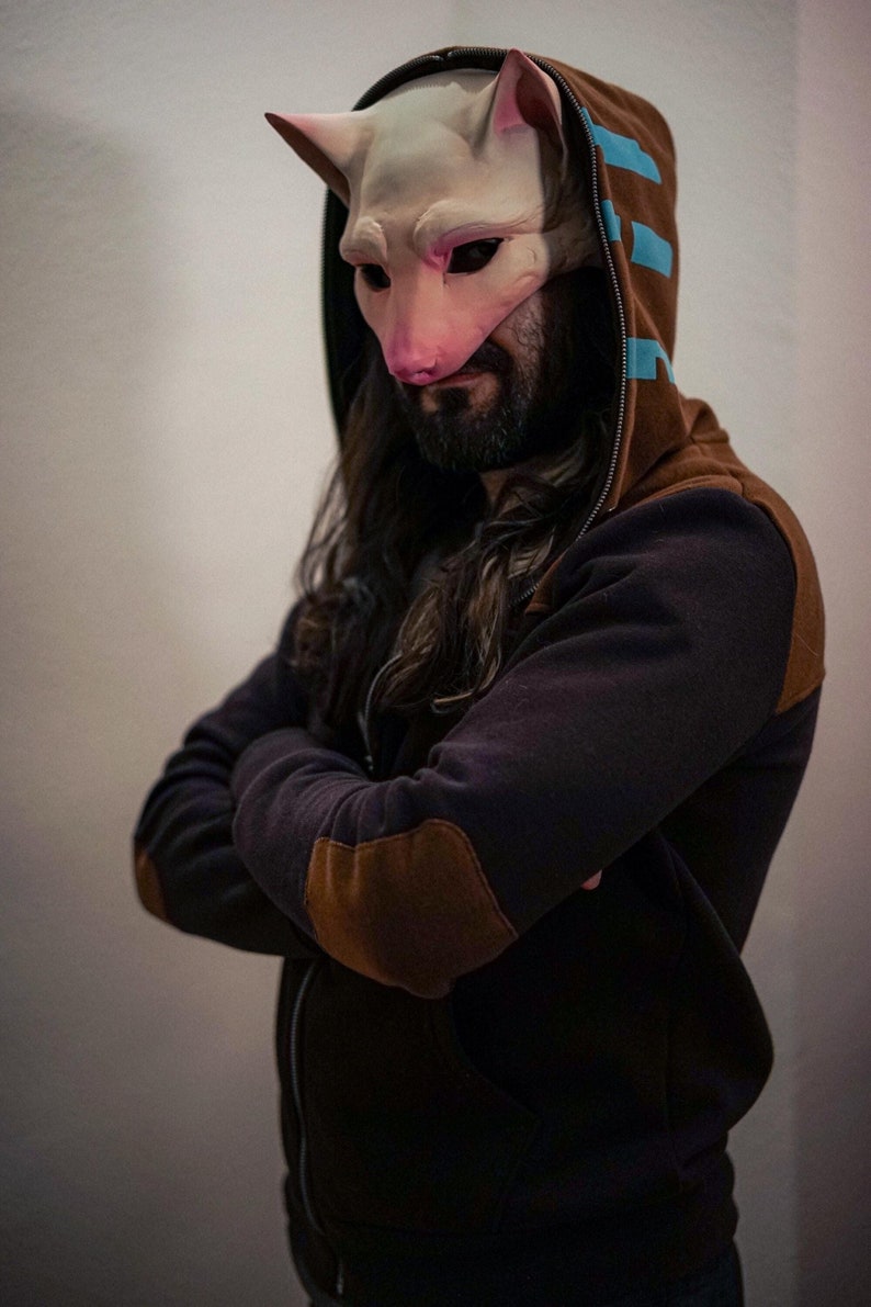 Op de afbeelding: Een persoon draagt een zwarte hoodie met bruine elleboogpatches en een wit kattenmasker met roze accenten.