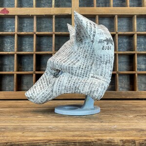 Op de afbeelding: Een sculptuur van een wolvenkop gemaakt van krantenpapier, gemonteerd op een lichtblauwe basis. De wolvenkop is naar links gericht, waarbij de textuur van het papier en de details van de vorm worden getoond. De achtergrond is een houten kast.