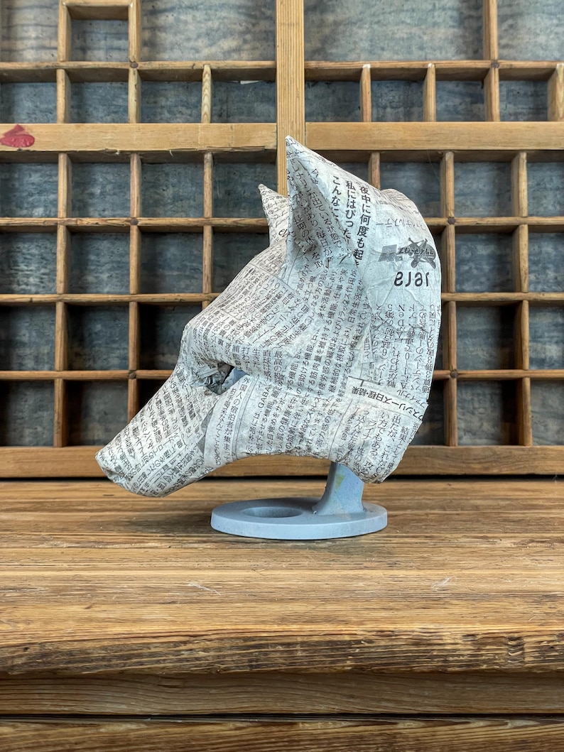Op de afbeelding: Een sculptuur van een wolvenkop gemaakt van krantenpapier, gemonteerd op een lichtblauwe basis. De wolvenkop is naar links gericht, waarbij de textuur van het papier en de details van de vorm worden getoond. De achtergrond is een houten kast.